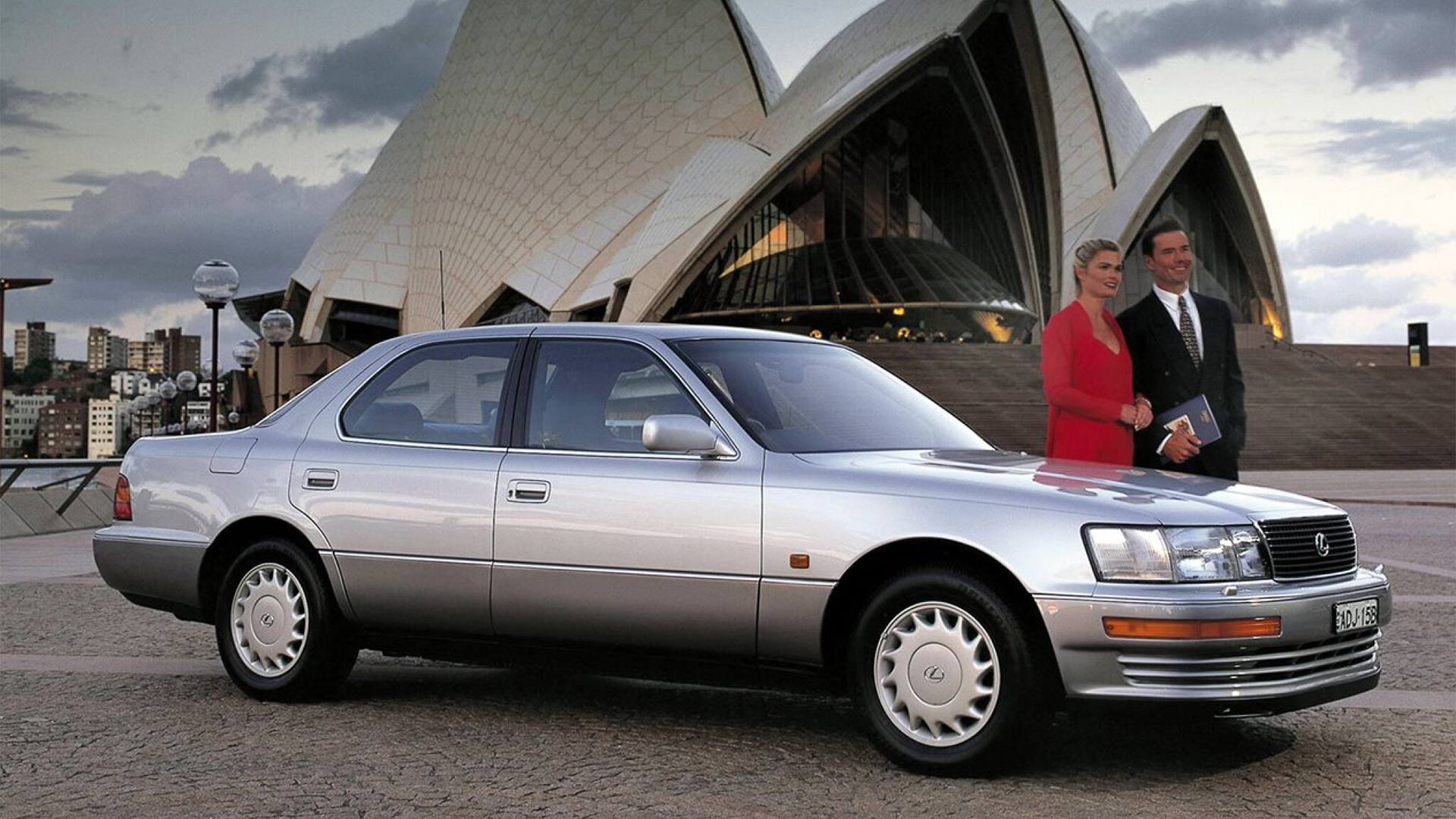 A man and woman stand with the first Lexus, silver,  in front of the Sydney Opera House. 