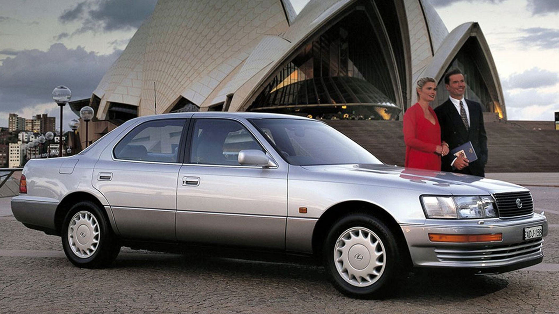 A silver Lexus vehicle parked next to two people in front of the Sydney Opera House