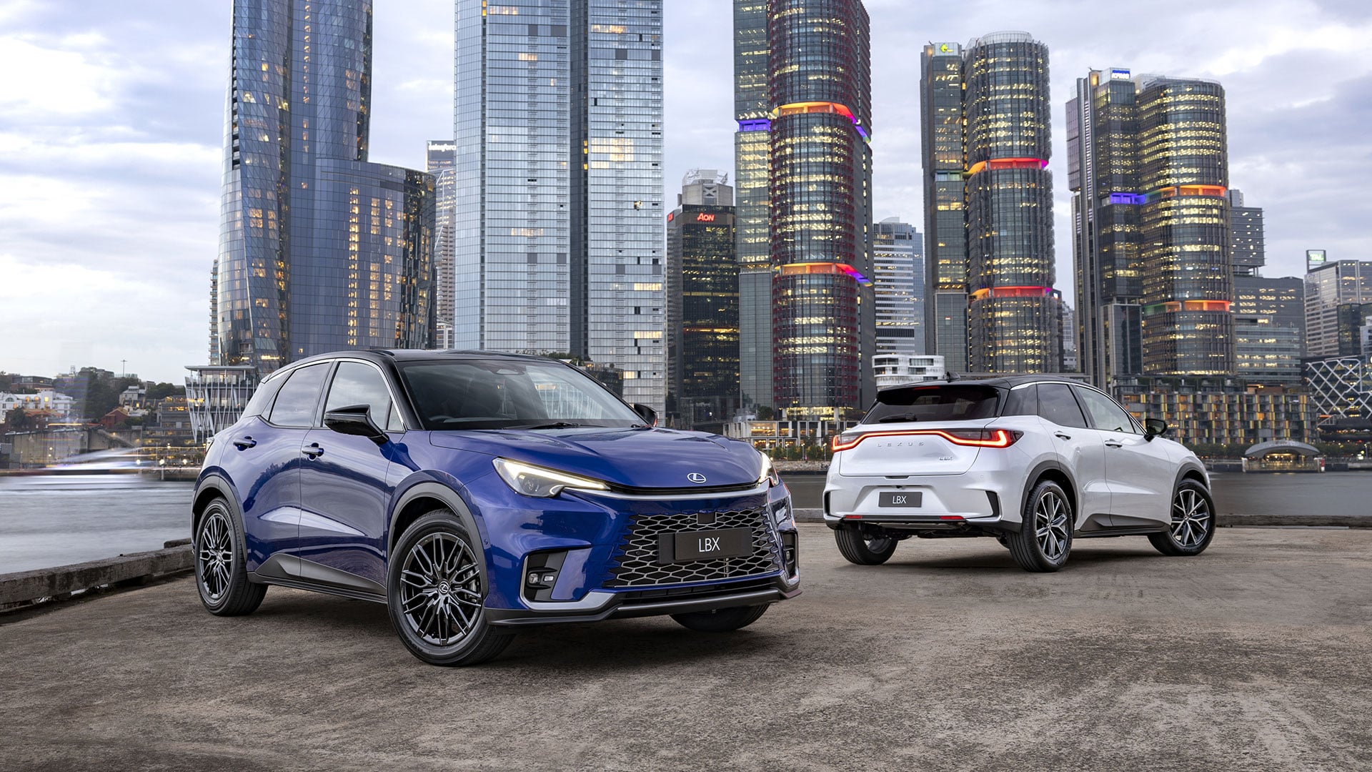 Two Lexus LBX vehicles, blue and white, parked in front of a city skyline