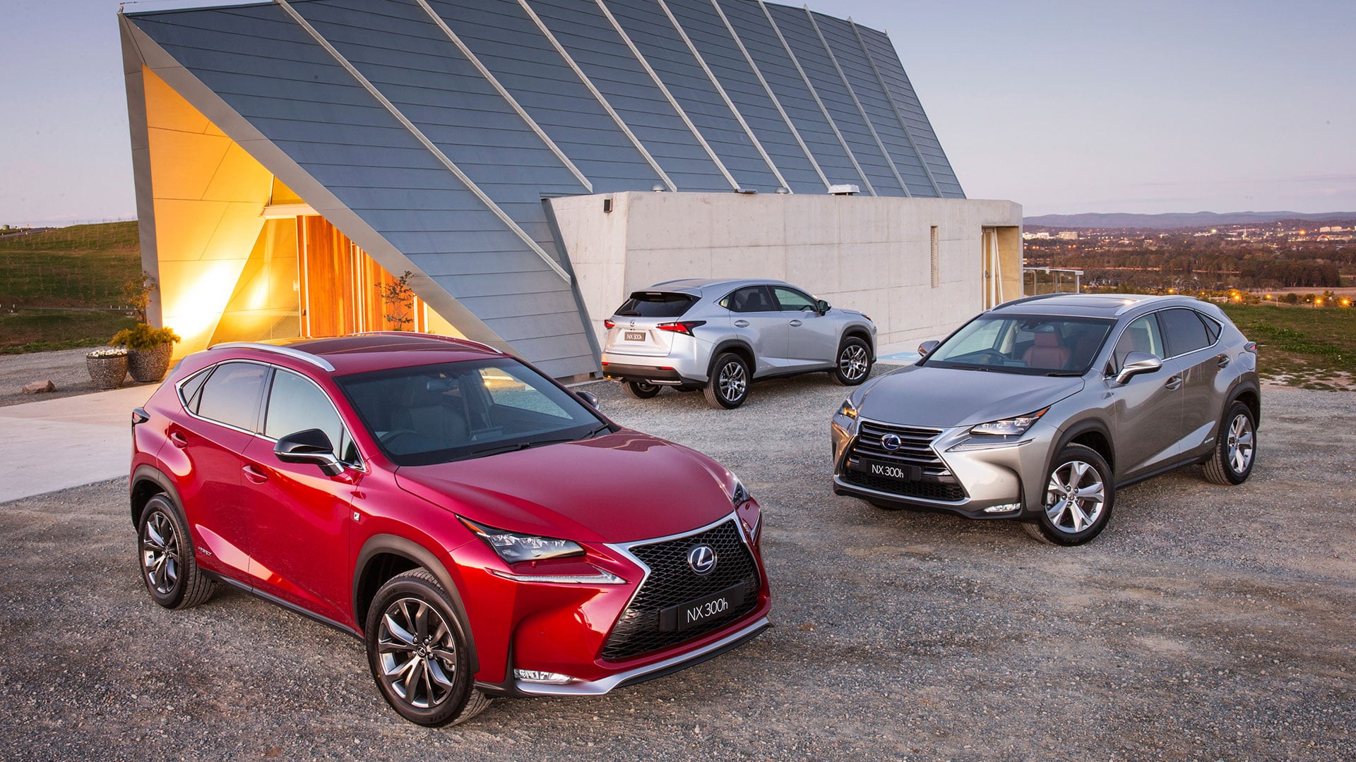 Three Lexus NX SUVs (red, silver, silver) parked in front of a modern building.
