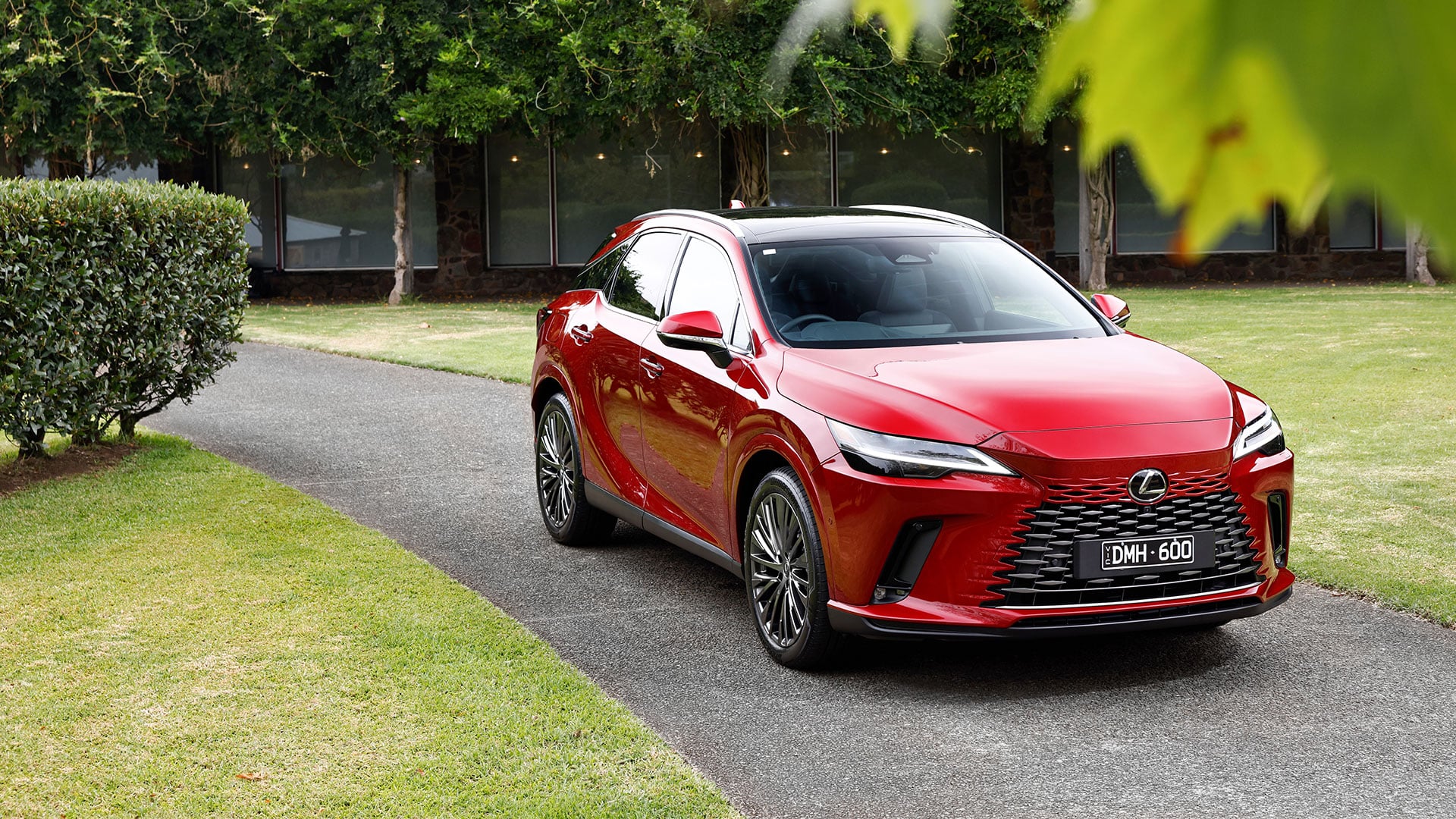 Red Lexus RX 450h+ plug-in hybrid luxury SUV parked on a paved driveway next to a grassy area.